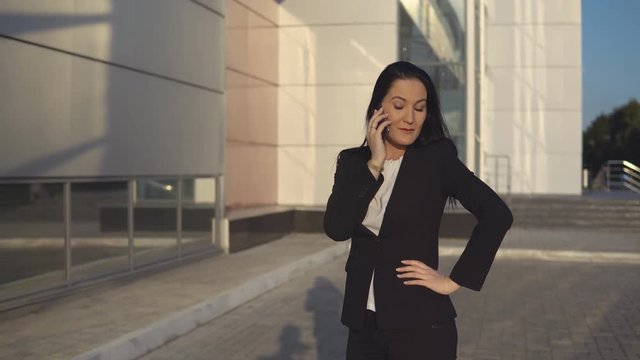 Businesswoman in formal suit talking on smartphone on sunny day against the backdrop of office center