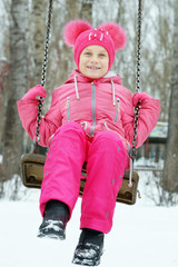 Little girl wearing bright clothes is swinging on a swing outdoors in winter park