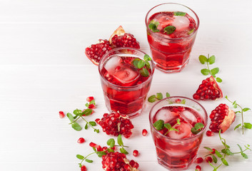 Pomegranate cocktail with ice and mint in beautiful glasses and jug, fresh ripe pomegranate on white wooden background. Sweet red juice. Close up photography. Selective focus