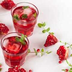Pomegranate cocktail with ice and mint in beautiful glasses and jug, fresh ripe pomegranate on white wooden background. Sweet red juice. Close up photography. Selective focus