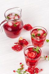 Pomegranate cocktail with ice and mint in beautiful glasses and jug, fresh ripe pomegranate on white wooden background. Sweet red juice. Close up photography. Selective focus