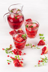 Pomegranate cocktail with ice and mint in beautiful glasses and jug, fresh ripe pomegranate on white wooden background. Sweet red juice. Close up photography. Selective focus