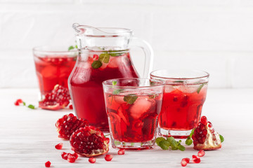 Pomegranate cocktail with ice and mint in beautiful glasses and jug, fresh ripe pomegranate on white wooden background. Sweet red juice. Close up photography. Selective focus