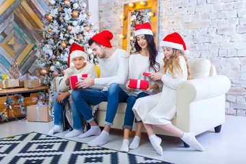 A Christmas photograph of a beautiful family with two children in Santa Claus red caps and red presents. Sit on the couch in the Christmas interior.