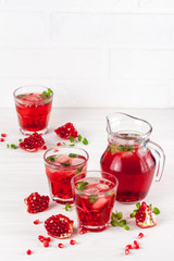 Pomegranate cocktail with ice and mint in beautiful glasses and jug, fresh ripe pomegranate on white wooden background. Sweet red juice. Close up photography. Selective focus