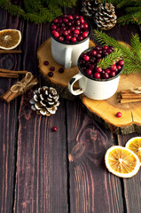Fresh red cranberries in a white cup, cinnamon sticks, dried orange circles, spruce branches and cones on a dark wooden background. Christmas background with berries.