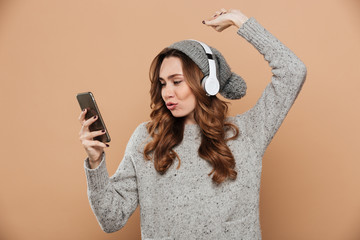 Cute young woman with curly hairstyle holding smartphone while listening to music