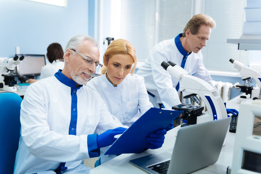 Making a research. Smiling delighted grey-haired scientist wearing glasses and showing something to a beautiful blond young woman and a serious biologist standing in the background