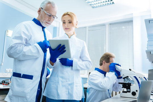 Analysing work. Grey-haired wrinkled researcher discussing genetics with a pretty blond young scientist while being in the lab and another researcher working with a microscope in the background