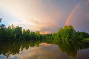 Rainbow over calm water of a forest lake. Beautiful clouds in the evening sky.