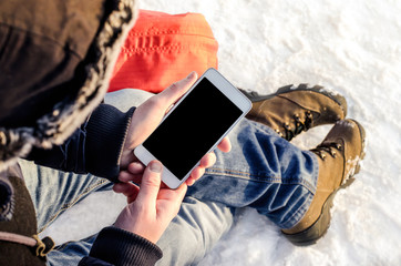 Hipster business man sitting in snow using white mobile smart phone, oled display, on wild nature, winter vacation, hiking, traveling, backpacker, warm clothes in Christmas time on a cold winter day
