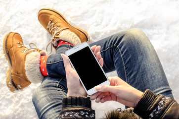 Hipster woman sitting in snow using white mobile smart phone, oled display, on wild nature, winter vacation, hiking, traveling, backpacker, warm clothes in Christmas time on a cold winter day