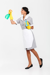 Full length photo of young brunette mais in uniform and yellow protective gloves spraying the cleaner on window