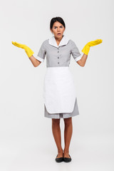 Full length photo of astonished maid in gray uniform and yellow rubber gloves standing with opened palms and looking at camera