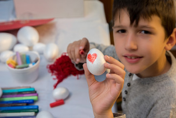 Young boy holding decorated Styrofoam egg  with red heart, surrounded with crufts and arts material, sitting at table 