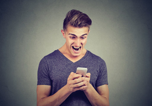 Frustrated angry man reading a text message on smartphone screaming isolated on gray background  