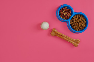 Dry pet food in bowl with a ball and dog bone on pink background top view