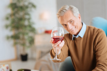 For you my darling. Selective focus on a glass of delicious red wine held by a cheerful retired gentleman sitting in a chair and smiling into the camera.