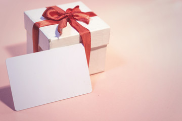 White gift box with red ribbon and white card on pink background