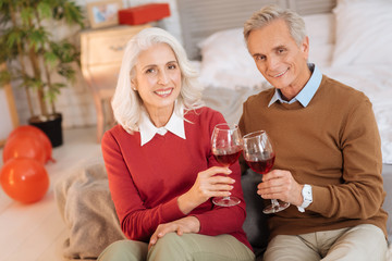 Family festivities. Cheerful senior couple looking into the camera with eyes full of happiness and smiling while enjoying red wine and celebrating anniversary together.