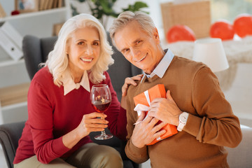 Harmonious marriage. Selective focus on a smiling retired gentleman holding a present while sitting next to his wife and celebrating their anniversary in a family circle.