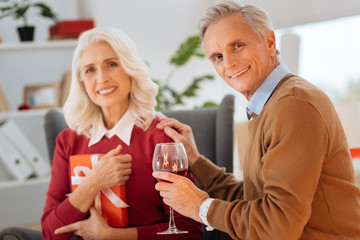 Dozens of years in love. Selective focus on a happy senior gentleman smiling into the camera and enjoying his glass of wine after surprising his wife with a gift.