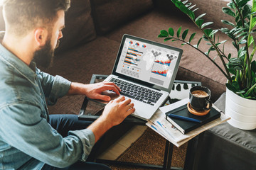 Young bearded hipster man, entrepreneur sits on couch at coffee table,uses laptop with graphs,charts, diagrams on screen.On table notebook,cup of coffee.Online marketing,education,e-learning. Startup.