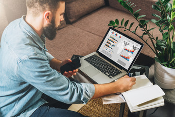 Man sits at table,working,holds smartphone,uses laptop with graphs, charts, diagrams on screen and makes notes in his notebook.Online marketing, education, e-learning, e-commerce. Startup, planning.