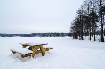 Wooden bench and table covered of snow in a snowy forest landscape. Frozen lake