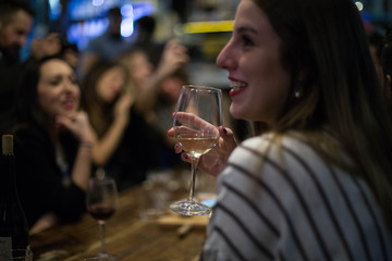 Young woman enjoys glass of dry white wine, chats and talks with friends at party or meeting in downtown alcohol bar, hipster beautiful going out style, open for communication and flirt