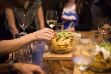 Group of friends hang out at downtown local cafe or bar at night, share party snack, bowl of mexican corn nachos topped with fresh guacamole. Partygoers eat tapas and drink wine