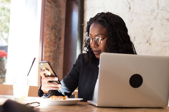 Beautiful african american woman with long hair and in prescription glasses uses her smartphone to chat about business deal or talk with friends, smiles with confidence and natural beauty