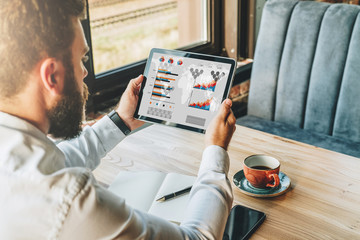 Back view. Young bearded businessman in white shirt is sitting at table, using tablet computer with graphs and charts, diagrams on screen, working, studying. Online marketing, education, e-learning