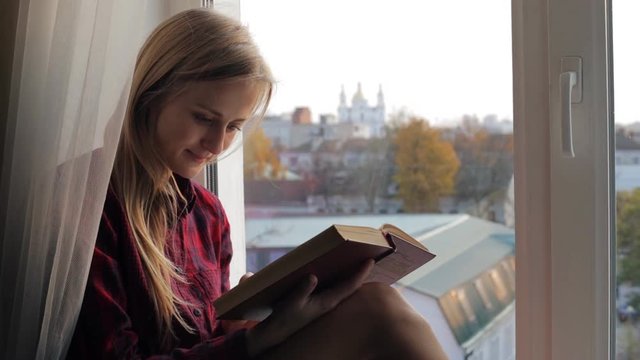 Beautiful blonde woman reading a book sitting on a windowsill at home
