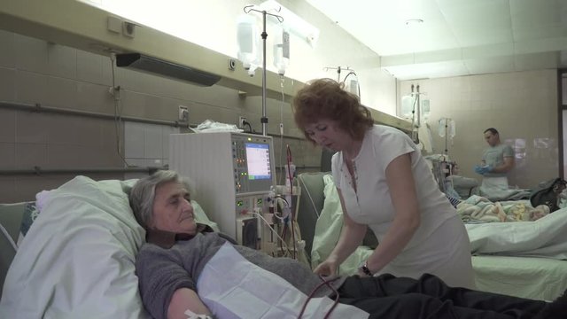 Group of hemodialysis mature female patients lying in the beds during peritoneal dialysis treatment at hospital room, nurse and male technician prepare machines and medicaments, steady cam shot