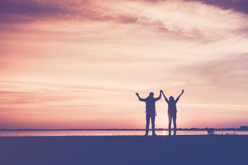 Silhouette of couple hikers with arms raised up and backpacks, relaxing and freedom between on walking on the Beach road at sunset. The concept of lovers and tourist traveler