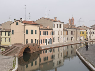 Fototapeta na wymiar Comacchio, FE, Italy - November 4, 2017: view of one of the canals crossing Comacchio. The colored facades of the houses are reflected on the water. On one side of the many bridges on the canal.