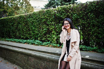 Black hair sexy woman in glasses and coat posed on street with mobile phone.