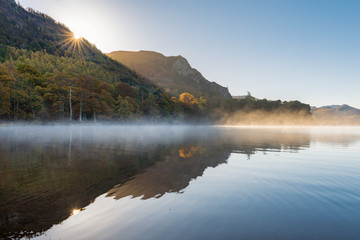 Naklejka premium Calm and peaceful misty Autumnal morning at Derwentwater in the Lake District as the sun rises above the mountains.