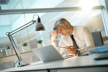Businessman in office having a headache in front of laptop - obrazy, fototapety, plakaty