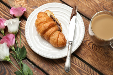 Plate with tasty croissant, flowers and coffee on wooden table