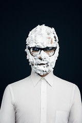 Closeup portrait of man with art makeup, beard, head in shaving foam with glasses in white shirt isolated at dark background