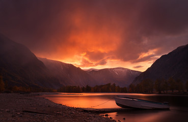 Sunset In The Mountains. Enchanting Autumn Mountain Landscape In Red Tones With Sunset Sky, River with Reflection And Lonely Boat. Mountain Valley With River And Boat On The Sunset Background. Altai