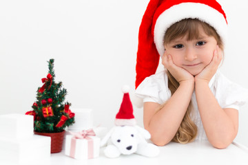 Little cute girl wearing a Santa Claus hat is sitting next to a little Christmas tree with a bunch of gifts and a plush dog. Christmas and New Year card. 2018 year.