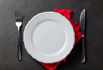 Festive set of knife and fork and white plate on a red napkin on a dark stone slate background, top view, copyspace