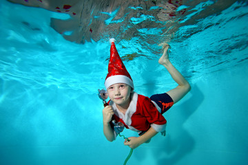 Obraz na płótnie Canvas A little boy in a cap and suit of Santa Claus swimming underwater with the phone in hand on blue background. Portrait. Shooting under water. Horizontal orientation
