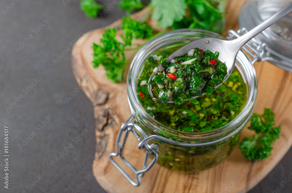 Wall mural Traditional argentinian chimichurri sauce made of parsley, cilantro, garlic and chili pepper in a glass jar, focus on a spoon with sauce. Selective focus, horizontal image