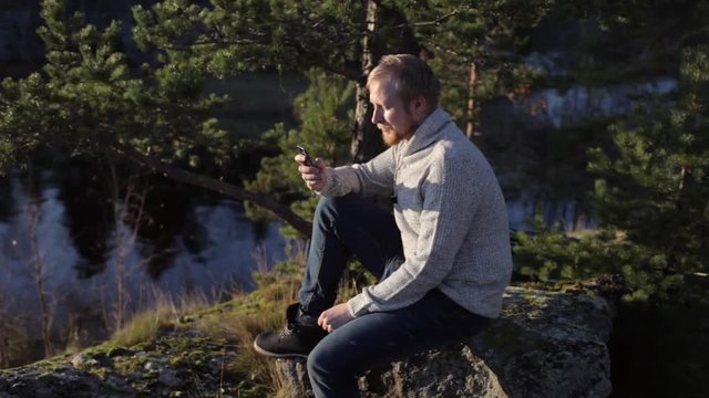 a man with a phone sits on the rocky shore of the lake