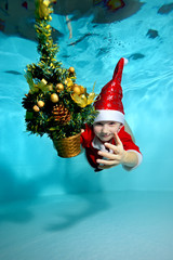 Child in costume Santa Claus swimming underwater with a Christmas tree in hand over blue background...