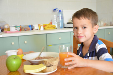 Cute boy drinking orange juice and eating muesli for breakfast. a healthy food on the table in the kitchen. Diet. morning. Plate with porridge, egg on a stand, bread, cheese, green apple. Carrot